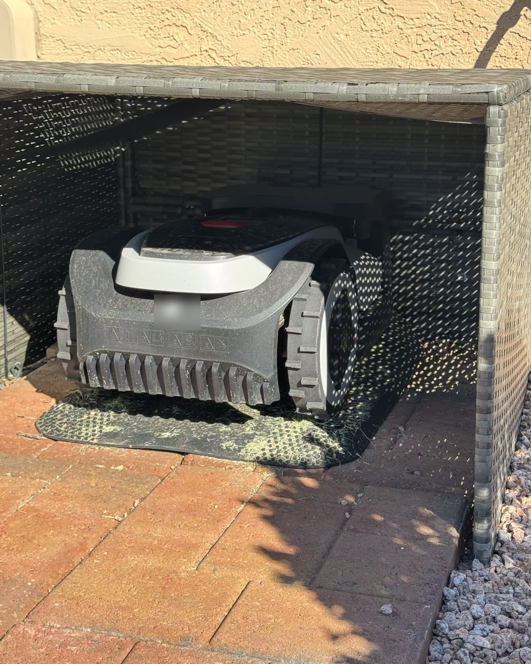 The robot mower parked on its charging mat inside a small woven shelter at the end of a paver path